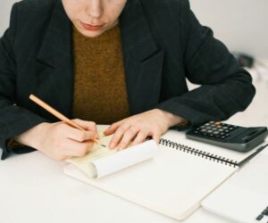 Person writing on a notepad at a desk with a calculator